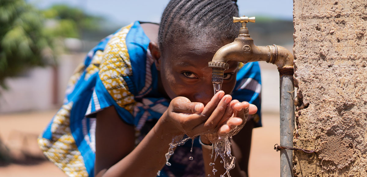 an image of a dark skinned woman drinking water from a tap attached to the outside of a brick building
