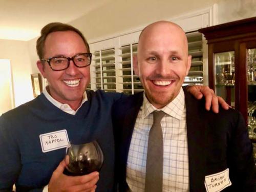 Two men with name tags and wine glasses smile at the camera